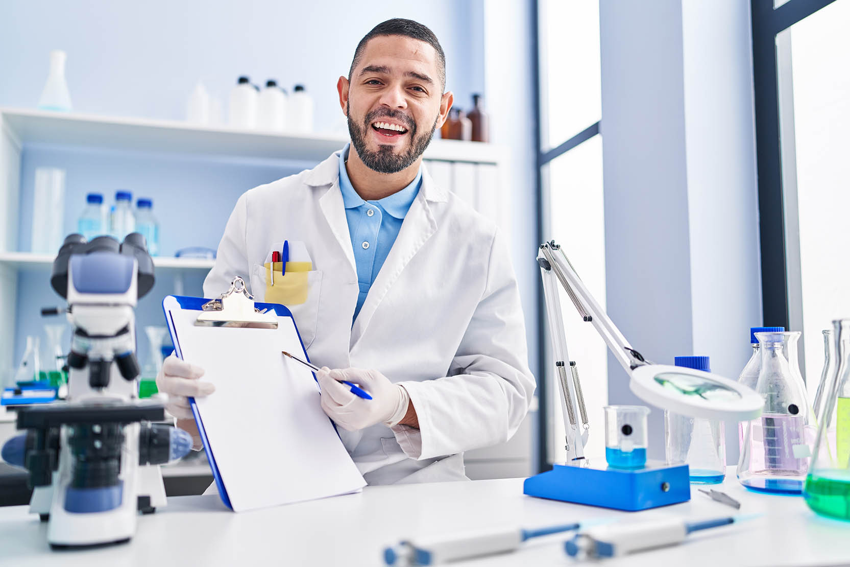 Hispanic,Man,Working,At,Scientist,Laboratory,Holding,Blank,Clipboard,Smiling
